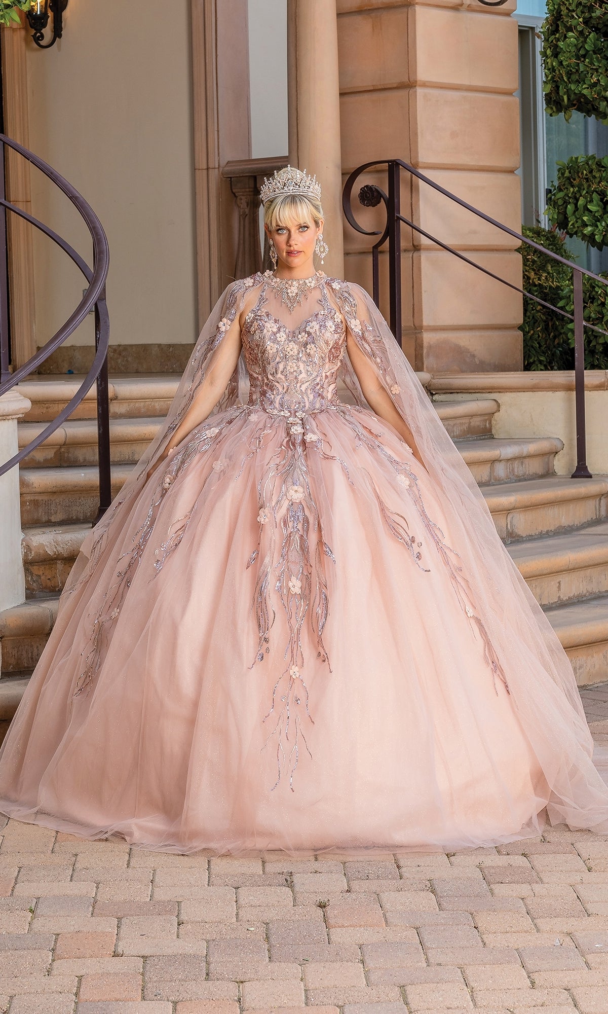 A person wears the Sleeveless Quinceanera Dress 1677, a pale pink ball gown with intricate floral embroidery and a removable sheer cape, paired with a jeweled crown, standing on stone steps in front of a building.